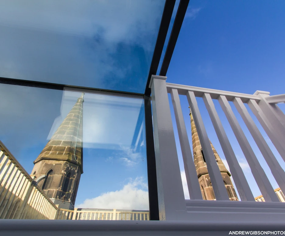 Reflection of a church tower in glass panels, framed by a white railing and a bright blue sky.