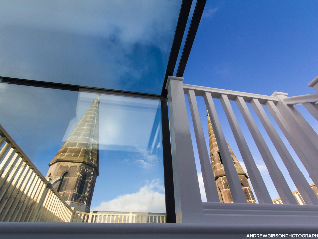 A glass structure reflects a tall church spire against a blue sky, with a white railing in the foreground creating a modern contrast to the historic architecture.