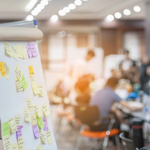 A flip chart covered with colorful sticky notes stands in the foreground, while a group of people is engaged in a collaborative activity in a blurred background. The setting appears to be a workshop or meeting room.