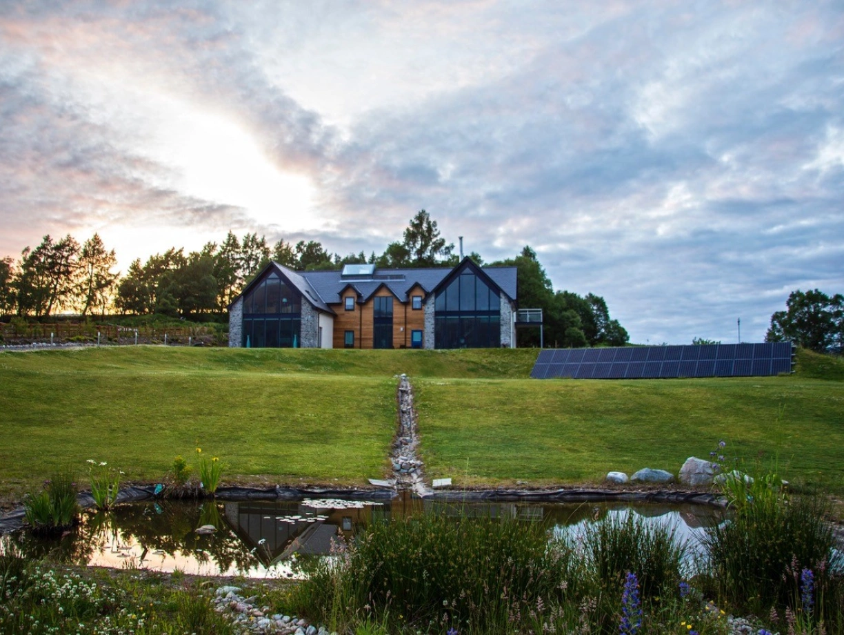 A modern house with large windows and a combination of wood and stone exterior sits on a grassy hillside. In the foreground, a small pond with surrounding plants reflects the cloudy sky at sunset. Solar panels are installed on the ground to the right of the house.