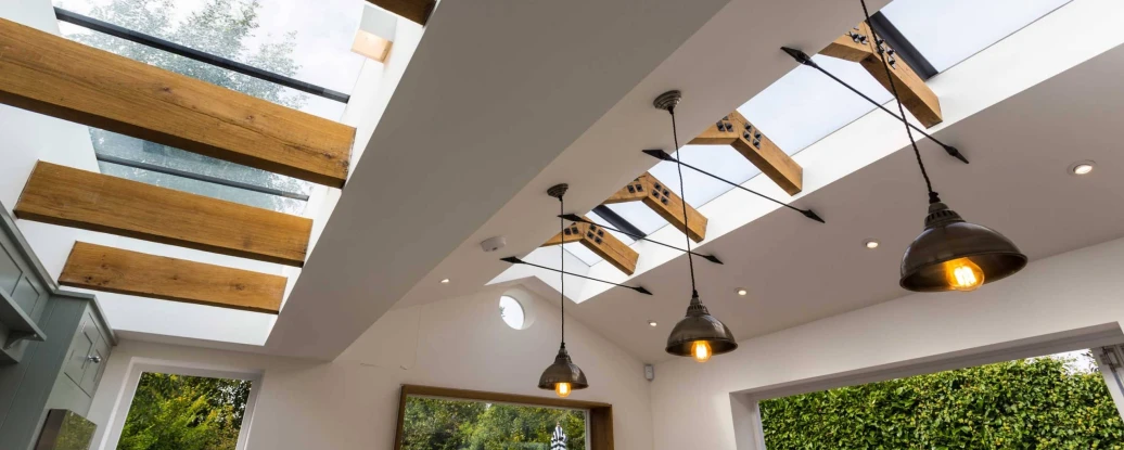 Modern interior featuring a bright room with skylights and wooden beams. Elegant light fixtures hang from the ceiling, and lush greenery is visible through a large window.