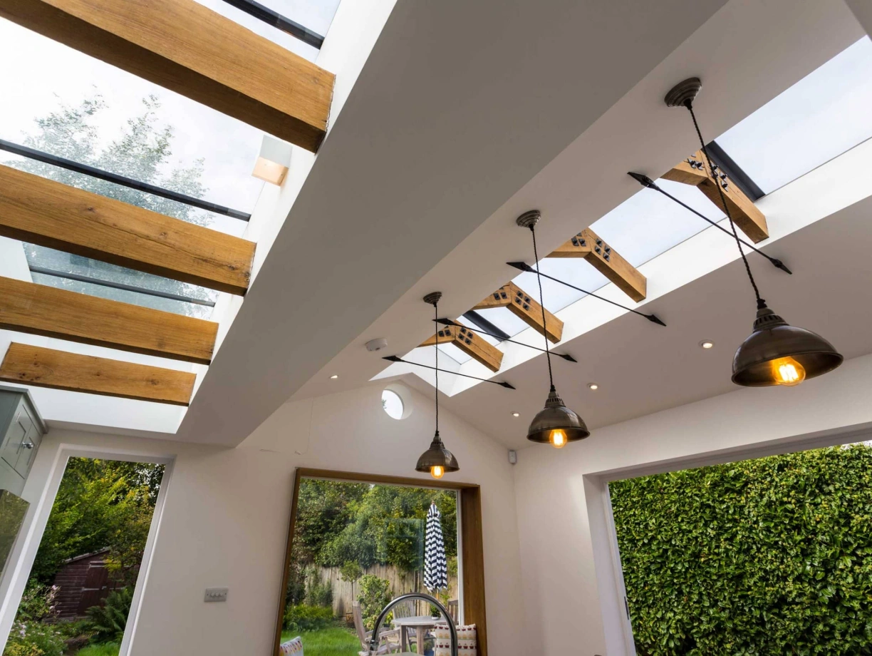Modern interior featuring a bright room with skylights and wooden beams. Elegant light fixtures hang from the ceiling, and lush greenery is visible through a large window.