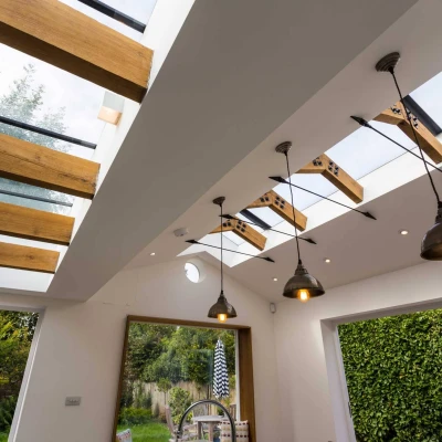 Modern interior featuring a bright room with skylights and wooden beams. Elegant light fixtures hang from the ceiling, and lush greenery is visible through a large window.