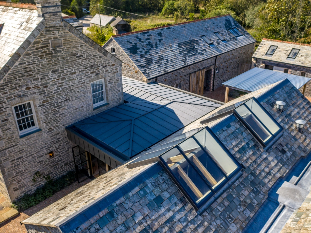 An aerial view of a rural property featuring a traditional stone house and several adjacent buildings. The roofs are made of slate and metal, with large skylights visible on one section. Surrounding greenery enhances the serene landscape.