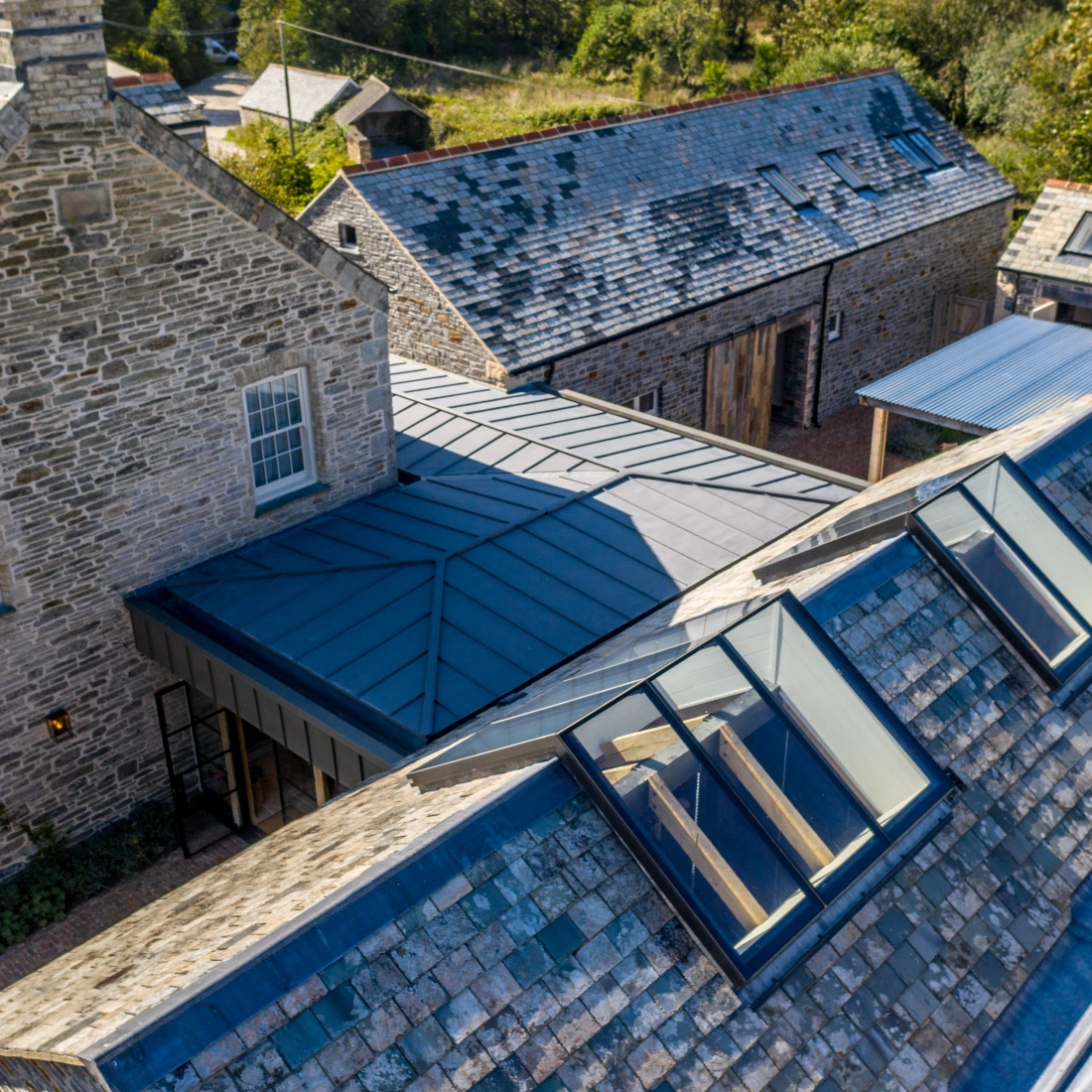 An aerial view of a rural property featuring a traditional stone house and several adjacent buildings. The roofs are made of slate and metal, with large skylights visible on one section. Surrounding greenery enhances the serene landscape.