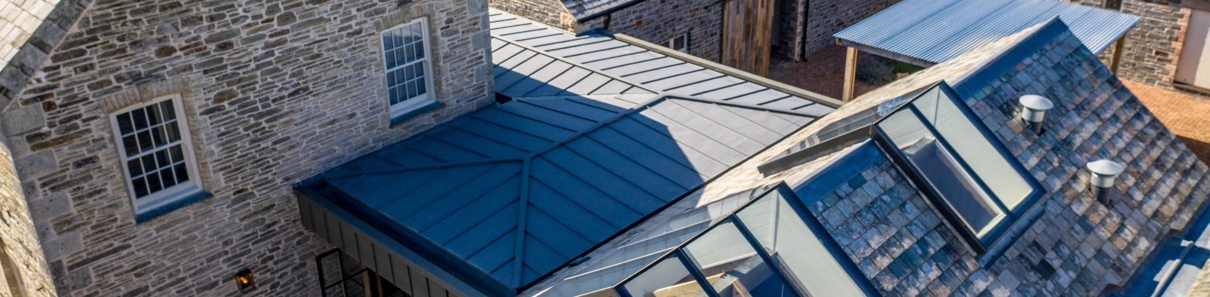 An aerial view of a rural property featuring a traditional stone house and several adjacent buildings. The roofs are made of slate and metal, with large skylights visible on one section. Surrounding greenery enhances the serene landscape.