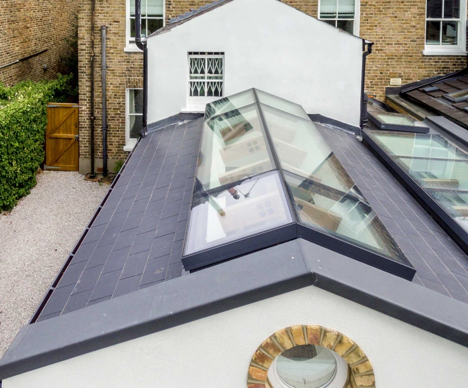 Aerial view of a modern flat roof with multiple glass skylights. Surrounding the roof are brick walls and a greenery-lined pathway leading to a wooden gate. The design features a blend of contemporary architecture with traditional elements.