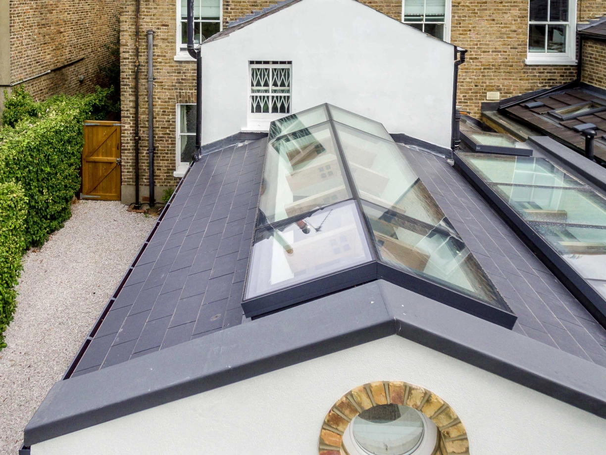 Aerial view of a modern flat roof with multiple glass skylights. Surrounding the roof are brick walls and a greenery-lined pathway leading to a wooden gate. The design features a blend of contemporary architecture with traditional elements.