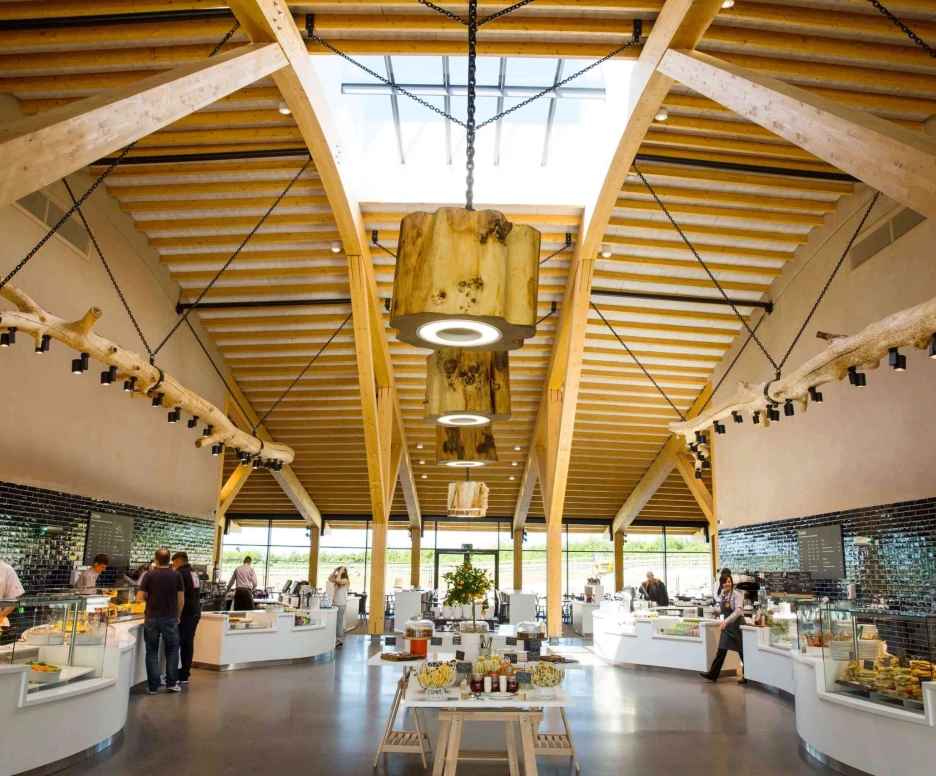A spacious, modern restaurant interior featuring high wooden ceilings and large windows. Several display counters showcase food items, while patrons and staff are engaged in conversation and service. Bright lighting highlights the open design and natural materials.