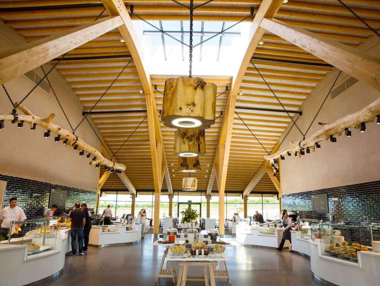 A spacious, modern restaurant interior featuring high wooden ceilings and large windows. Several display counters showcase food items, while patrons and staff are engaged in conversation and service. Bright lighting highlights the open design and natural materials.