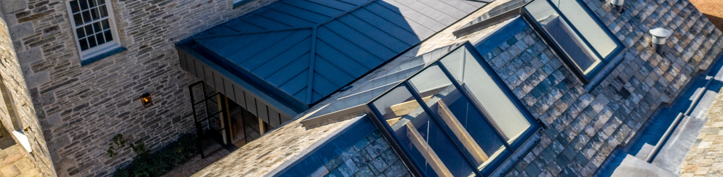 Vue aérienne d’un ensemble de bâtiments en pierre avec des toits en ardoise. Les structures comprennent une maison principale et plusieurs dépendances, toutes situées dans un paysage verdoyant. Des lucarnes en verre sont visibles sur un toit incliné.