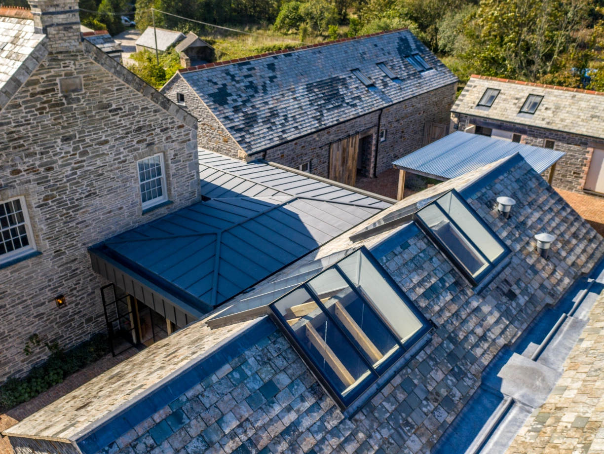 Vue aérienne d’un ensemble de bâtiments en pierre avec des toits en ardoise. Les structures comprennent une maison principale et plusieurs dépendances, toutes situées dans un paysage verdoyant. Des lucarnes en verre sont visibles sur un toit incliné.