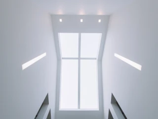 A view looking upward at a bright, minimalist stairwell. The ceiling is mostly white, featuring recessed lighting and a large rectangular window that lets in natural light.