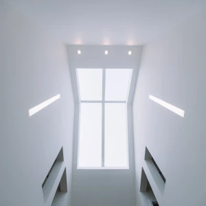A view looking upward at a bright, minimalist stairwell. The ceiling is mostly white, featuring recessed lighting and a large rectangular window that lets in natural light.