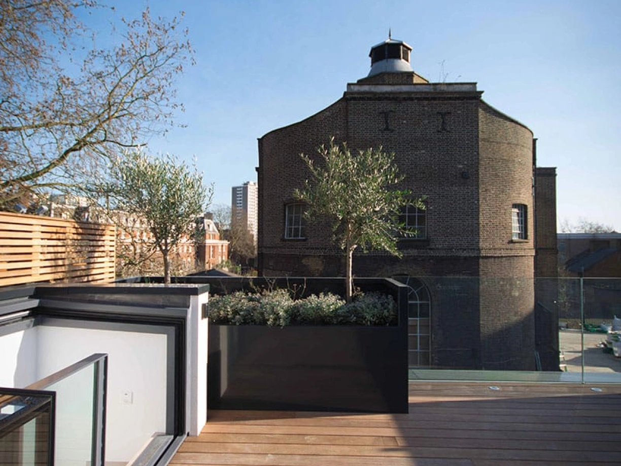 A rooftop terrace features wooden flooring and a large planter with small trees, overlooking a historic brick building with a round structure under a clear blue sky.