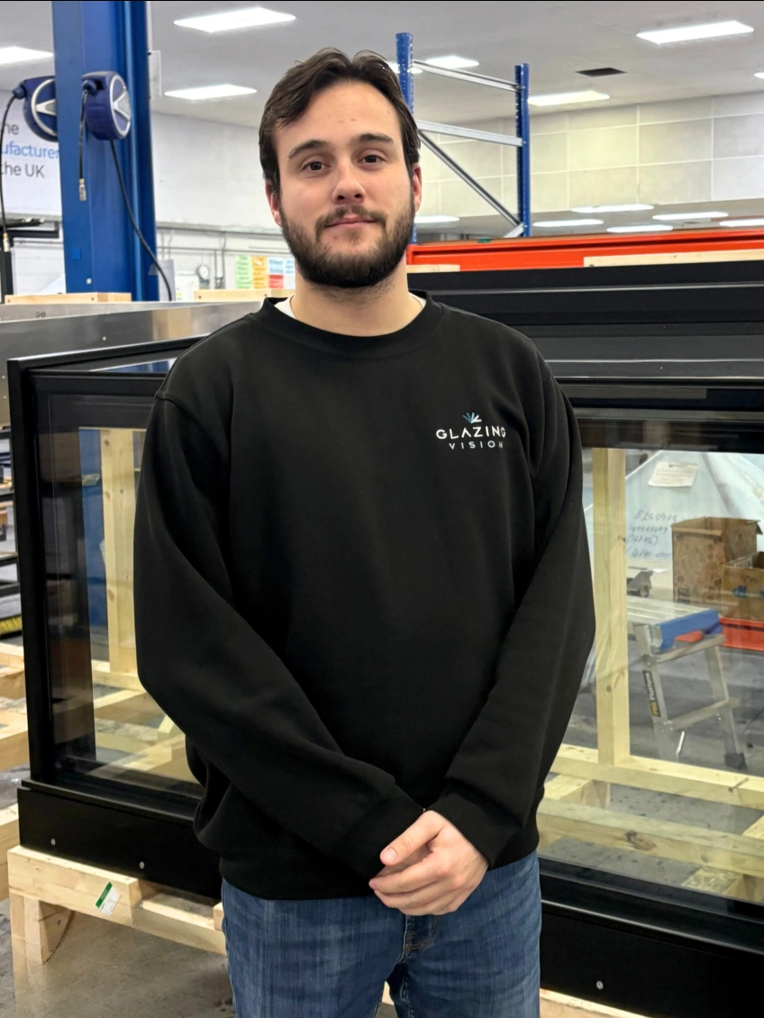 A young man in a black sweatshirt stands in front of a large glass display case in a workshop setting. He has brown hair and a slight smile. The background includes shelves and equipment typical of a manufacturing environment.
