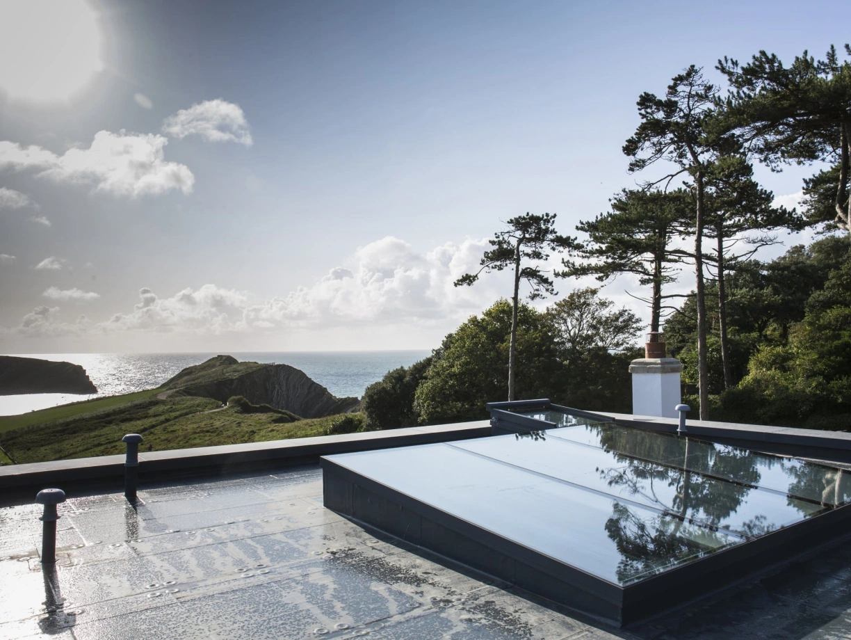 A rooftop view featuring skylights, overlooking a coastal landscape with cliffs, trees, and the ocean under a partly cloudy sky. The sun shines brightly in the background.