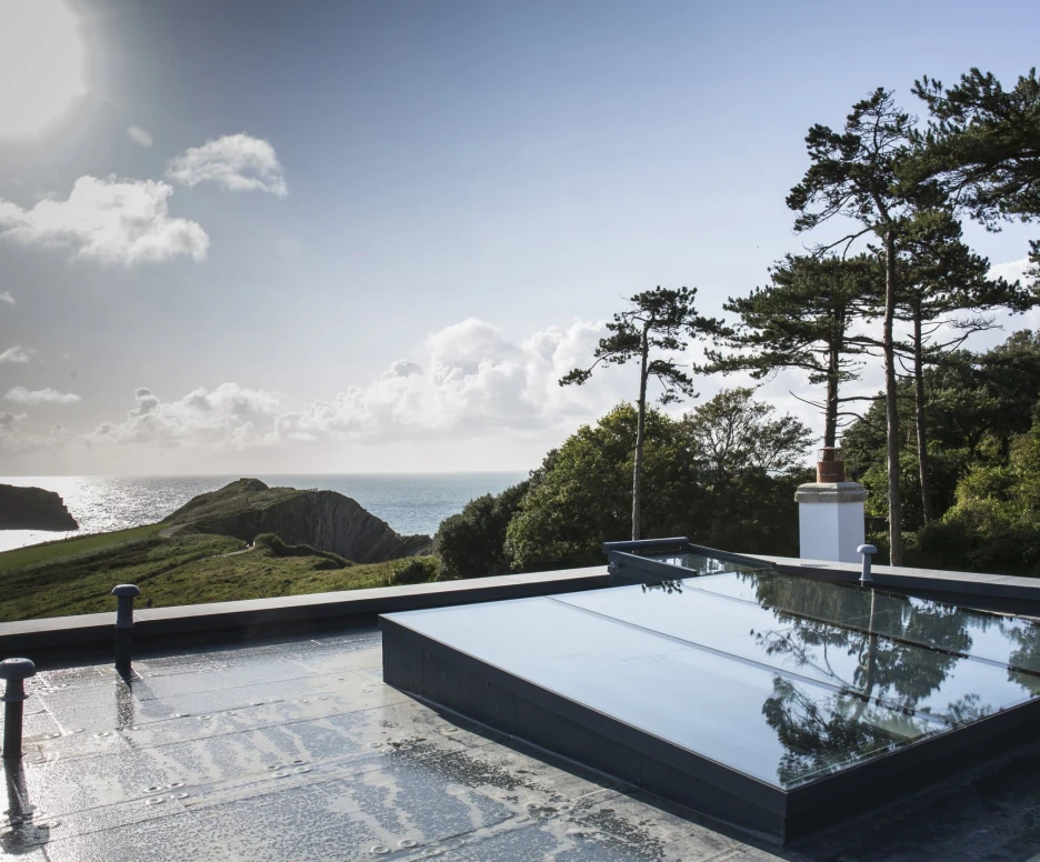 A rooftop view featuring skylights, overlooking a coastal landscape with cliffs, trees, and the ocean under a partly cloudy sky. The sun shines brightly in the background.