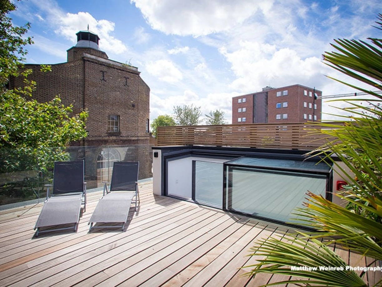 A modern rooftop deck features two lounge chairs, surrounded by greenery and sunlight. In the background, a historic brick building contrasts with a nearby contemporary apartment block under a blue sky with scattered clouds.
