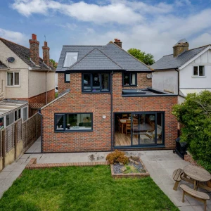 A modern two-story house made of red brick with large windows, surrounded by a well-maintained garden featuring a patio area and wooden seating. Neighbors' homes can be seen in the background under a partly cloudy sky.
