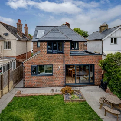 A modern two-story house made of red brick with large windows, surrounded by a well-maintained garden featuring a patio area and wooden seating. Neighbors' homes can be seen in the background under a partly cloudy sky.