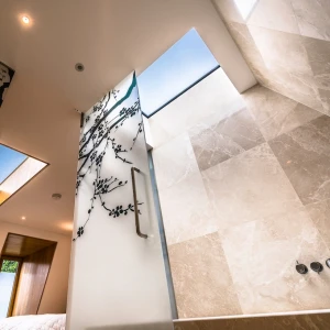 A modern bathroom with a sleek design featuring large stone tiles, a glass wall with artistic branch designs, and skylights allowing natural light to enter. The view is from below, highlighting the shower area and the visual harmony of the space.