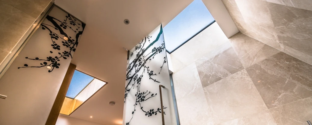 A modern bathroom featuring elegant stone walls, skylights, and decorative black floral patterns on a glass panel. Natural light illuminates the space, creating a bright and airy atmosphere.