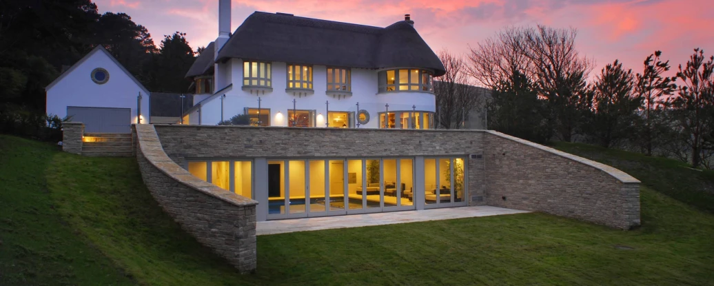 A modern house with a thatched roof, featuring large windows and a stone wall, set on a green hillside against a colorful sunset sky.