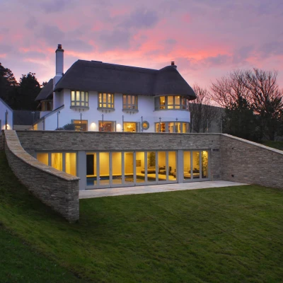 A modern house with a thatched roof, featuring large windows and a stone wall, set on a green hillside against a colorful sunset sky.
