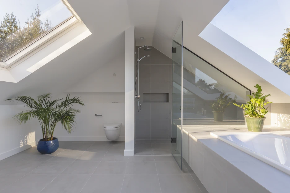A modern bathroom featuring sloped ceilings, a glass shower enclosure, and a large window. The space includes a toilet and a potted plant in a blue pot, along with another plant on the countertop beside a bathtub. Natural light floods the room.