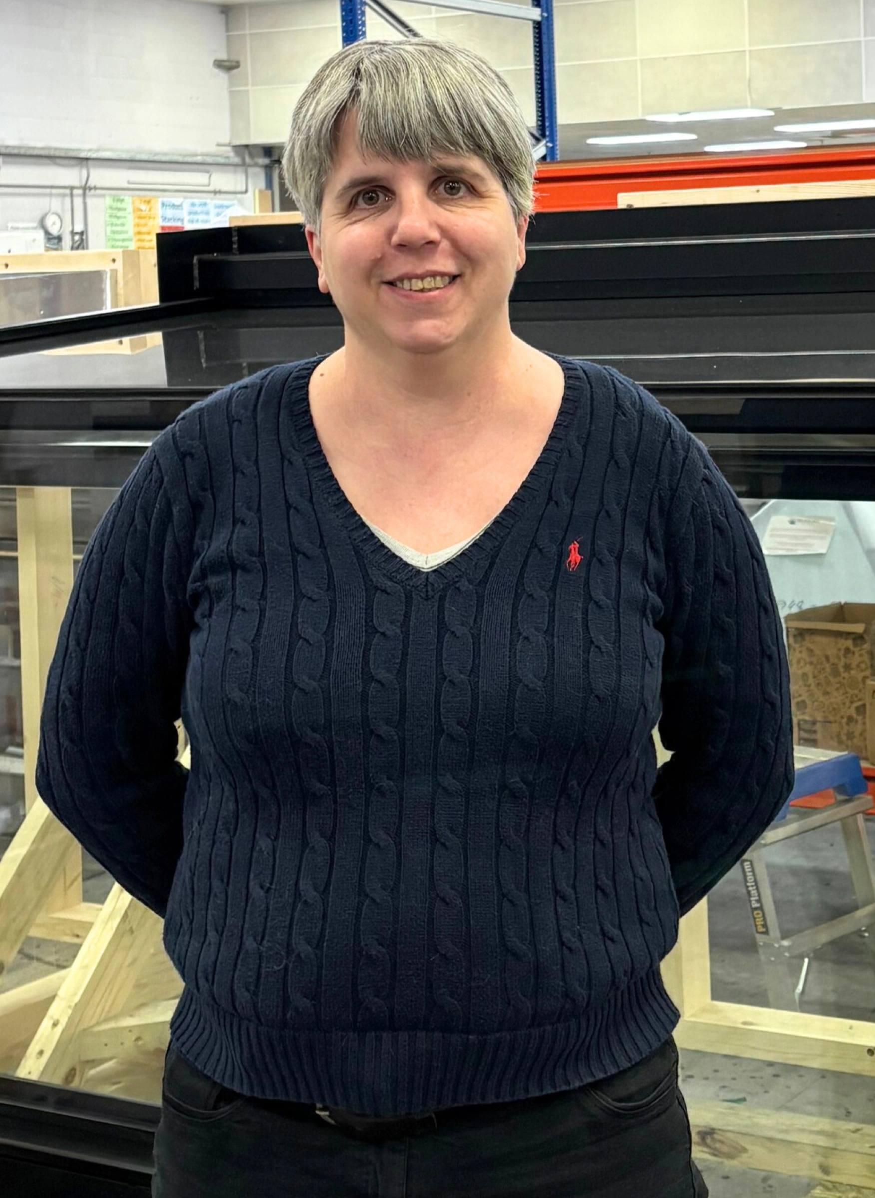 A woman with short gray hair smiles while standing in front of a large glass structure. She wears a navy blue cable-knit sweater and black pants.