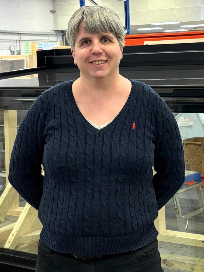A woman with short gray hair smiles while standing in front of a large glass structure. She wears a navy blue cable-knit sweater and black pants.