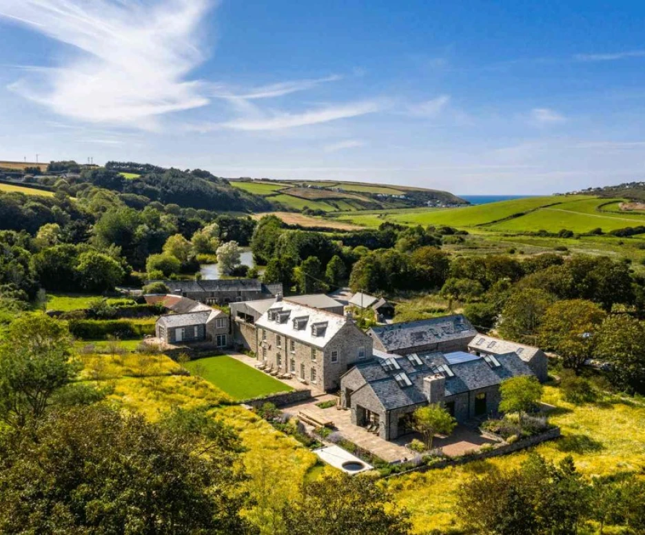 A picturesque rural landscape featuring a large stone house surrounded by lush green fields and trees, with a view of the sea in the distance under a clear blue sky.