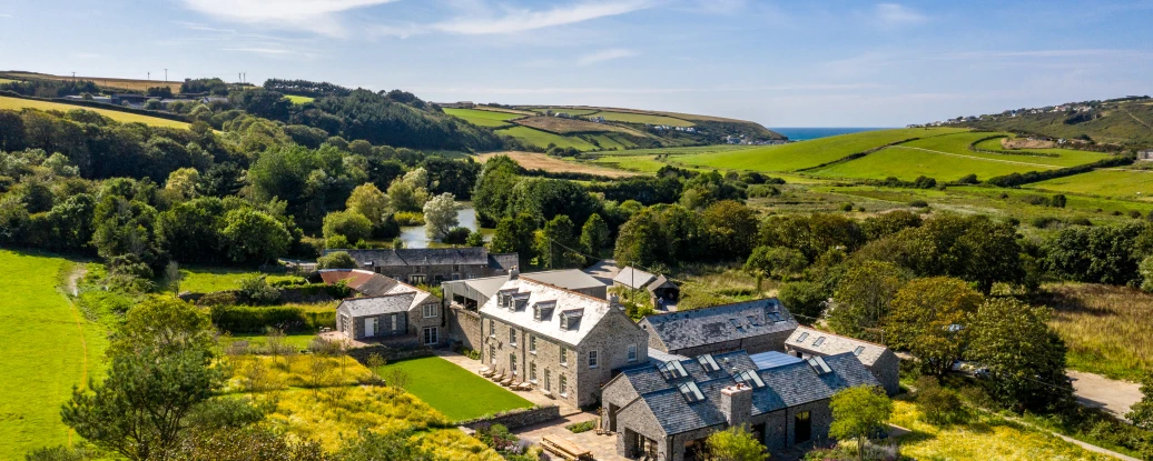 Aerial view of a large stone farmhouse surrounded by lush fields and trees, with rolling hills and the sea visible in the background under a clear blue sky.