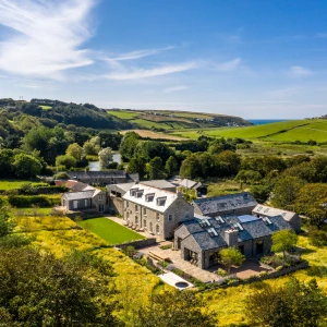 A picturesque countryside scene featuring a large stone house with multiple wings, surrounded by lush green fields and trees. Rolling hills extend into the distance, and the coastline is visible under a bright blue sky with wispy clouds.
