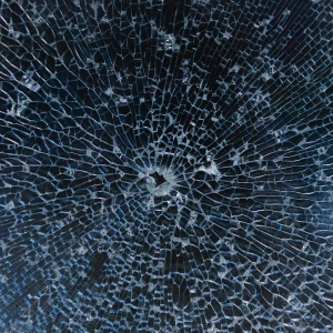 A close-up of a shattered glass surface, featuring a web-like pattern of cracks radiating from a central point. The glass has a shiny, reflective quality with varying shades of blue.