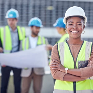 A confident woman wearing a safety helmet and a high-visibility vest stands in the foreground, smiling and crossing her arms. In the background, a group of people in hard hats and similar vests discuss plans and collaborate on a construction site.