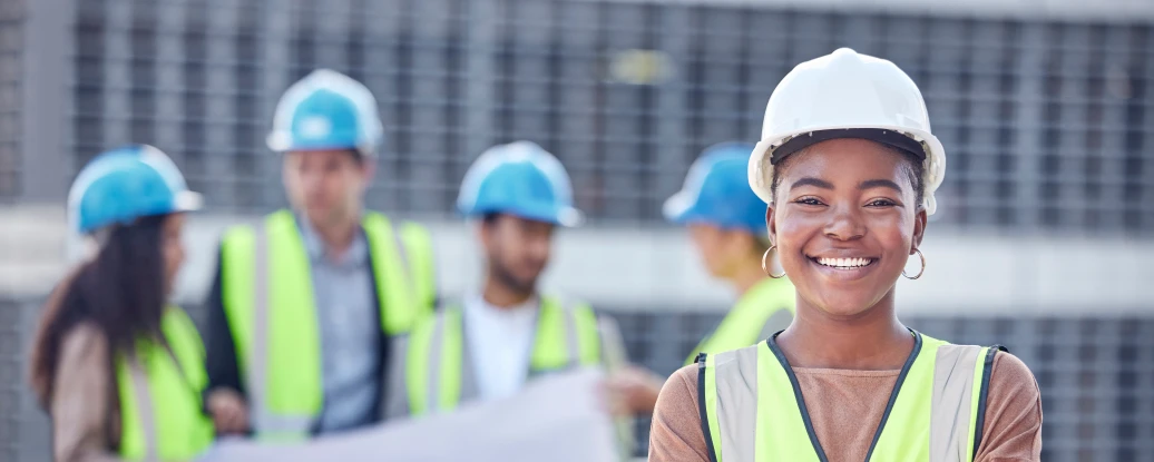 A confident woman wearing a safety helmet and a high-visibility vest stands in the foreground, smiling and crossing her arms. In the background, a group of people in hard hats and similar vests discuss plans and collaborate on a construction site.