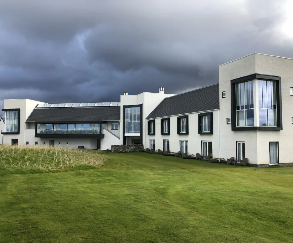 A modern white building with large windows, featuring a grassy lawn in front and dramatic grey clouds overhead.