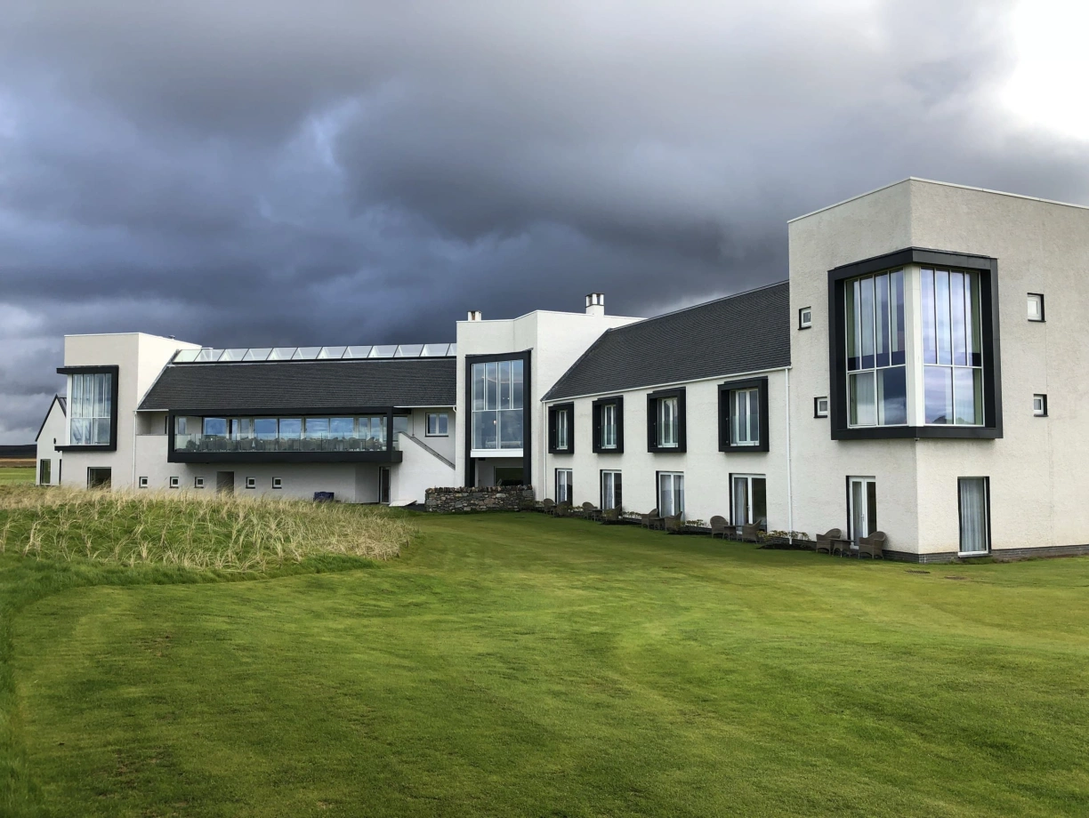 A modern white building with large windows, featuring a grassy lawn in front and dramatic grey clouds overhead.