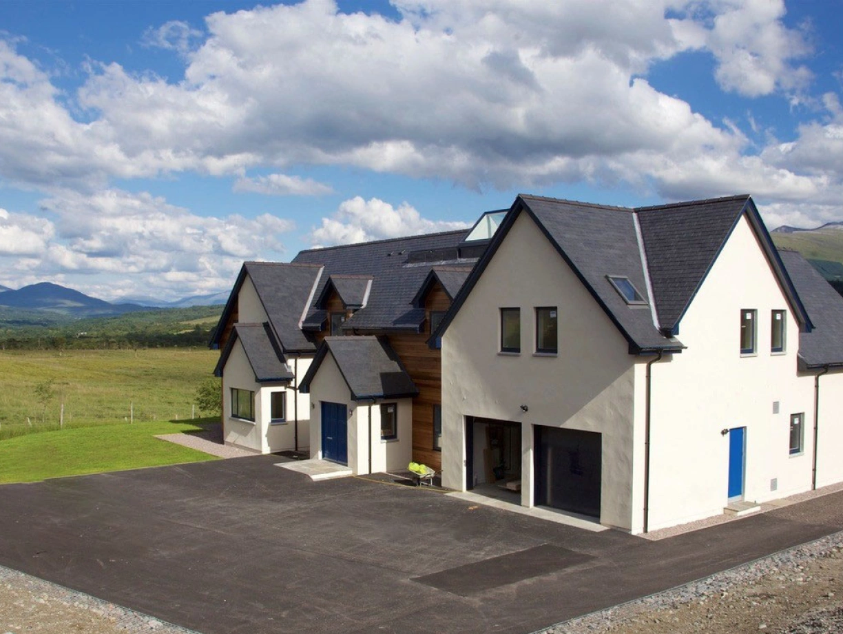 A modern two-story house with a sleek design, featuring a mix of white walls and wooden accents. Large windows and blue doors complement the exterior. The house is surrounded by a green lawn and open fields, under a bright blue sky with scattered clouds and distant mountains.