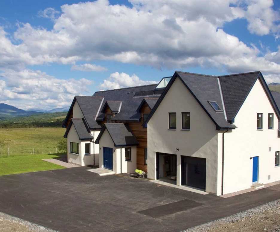 Modern two-story house with a mix of white and wooden exterior, situated in a scenic landscape. Lush green fields and mountains are visible in the background under a blue sky filled with fluffy clouds. A spacious driveway is in front of the house.