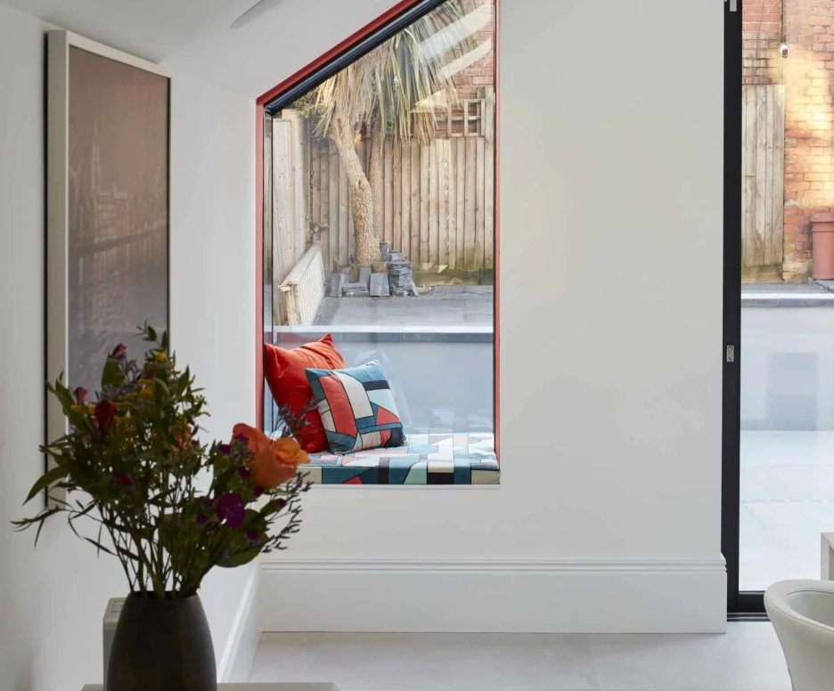 A bright, modern interior with a large window featuring a slanted design. Outside, a small outdoor space is visible, including a wooden fence and a palm tree. A vase of flowers sits on a table in the foreground, and colorful cushions are arranged in the window area.