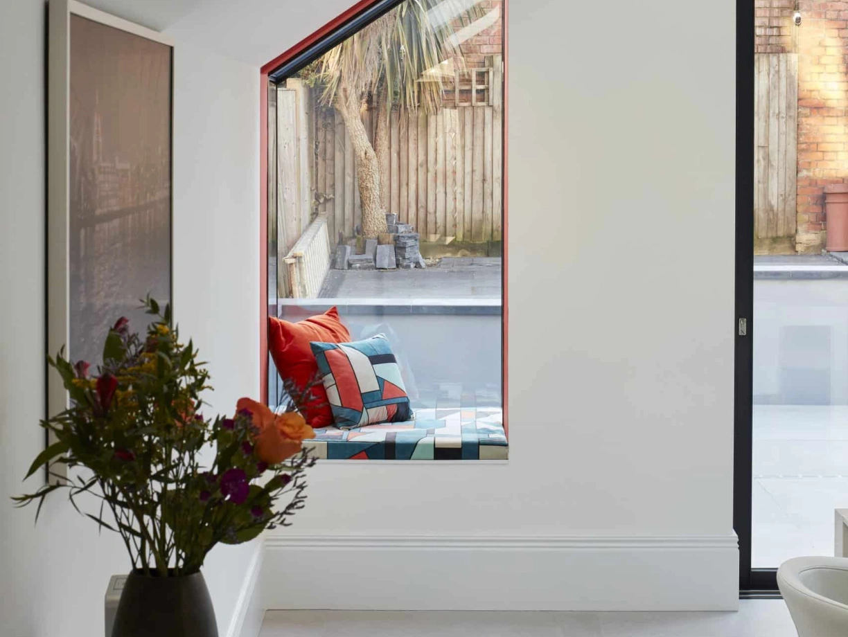 A bright, modern interior with a large window featuring a slanted design. Outside, a small outdoor space is visible, including a wooden fence and a palm tree. A vase of flowers sits on a table in the foreground, and colorful cushions are arranged in the window area.
