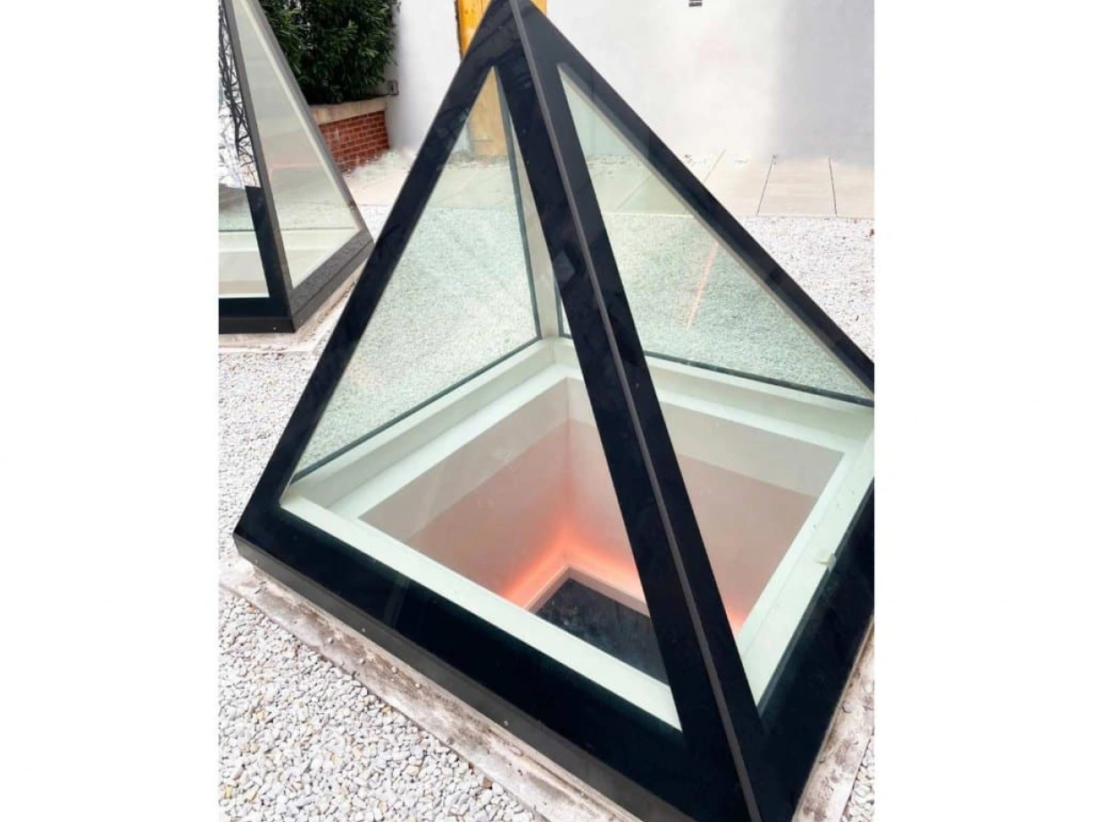 A modern glass pyramid with a black frame sits in a gravel area. The interior features soft, illuminated edges, creating a warm glow.