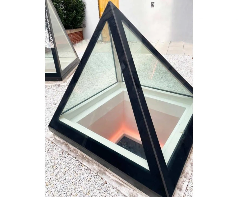 A modern glass pyramid with a black frame sits in a gravel area. The interior features soft, illuminated edges, creating a warm glow.