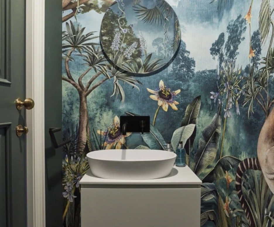 A stylish bathroom featuring a tropical mural with greenery and flowers. A round mirror hangs above a modern, white sink mounted on a minimalistic cabinet. Black and white checkered flooring adds a touch of elegance.