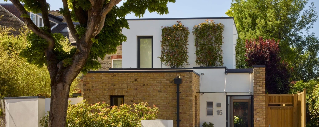 Modern two-story house with a combination of brick and white walls. The upper floor features windows adorned with green plants, while the lower floor has a neatly landscaped garden and a wooden gate. Lush trees provide a natural backdrop.
