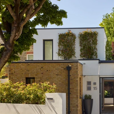 Modern two-story house with a combination of brick and white walls. The upper floor features windows adorned with green plants, while the lower floor has a neatly landscaped garden and a wooden gate. Lush trees provide a natural backdrop.
