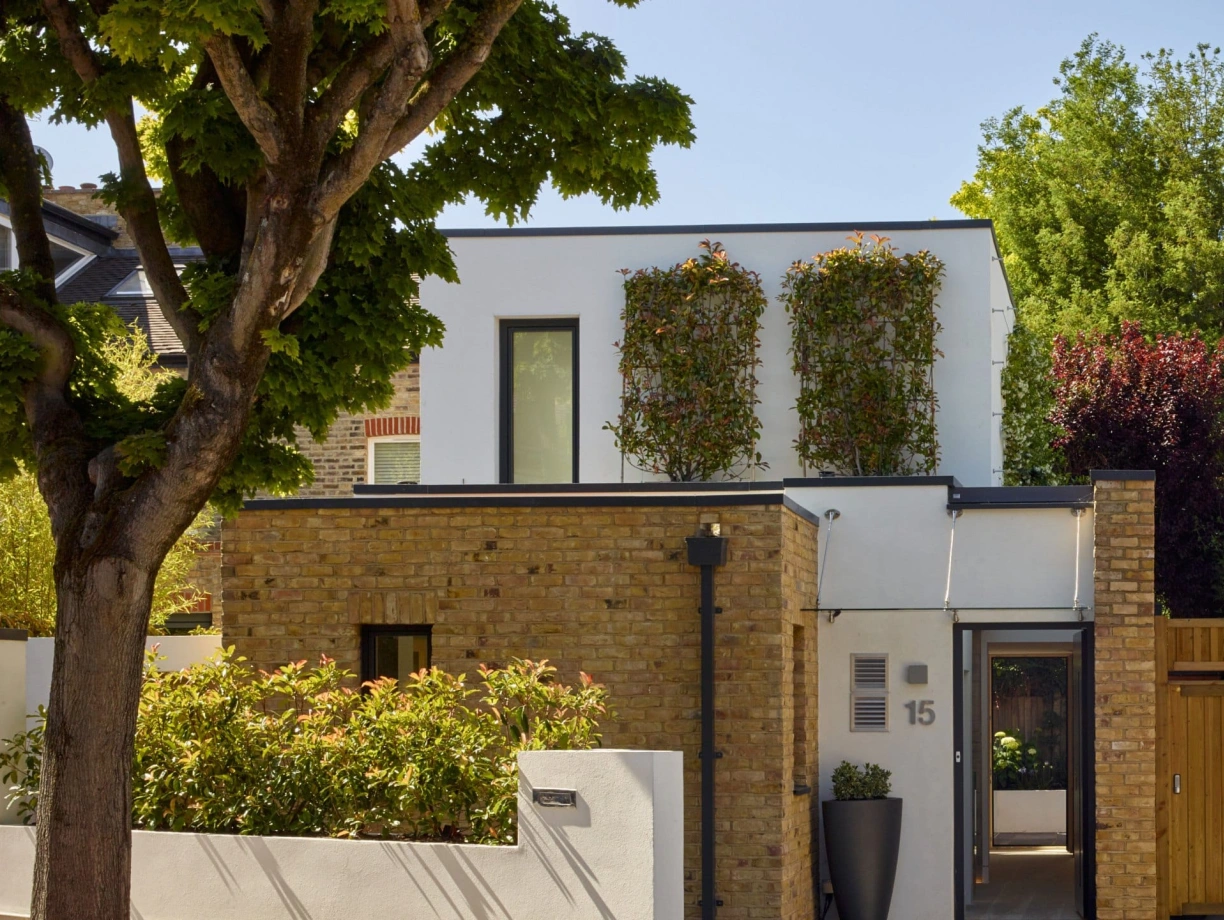 Modern two-story house with a combination of brick and white walls. The upper floor features windows adorned with green plants, while the lower floor has a neatly landscaped garden and a wooden gate. Lush trees provide a natural backdrop.